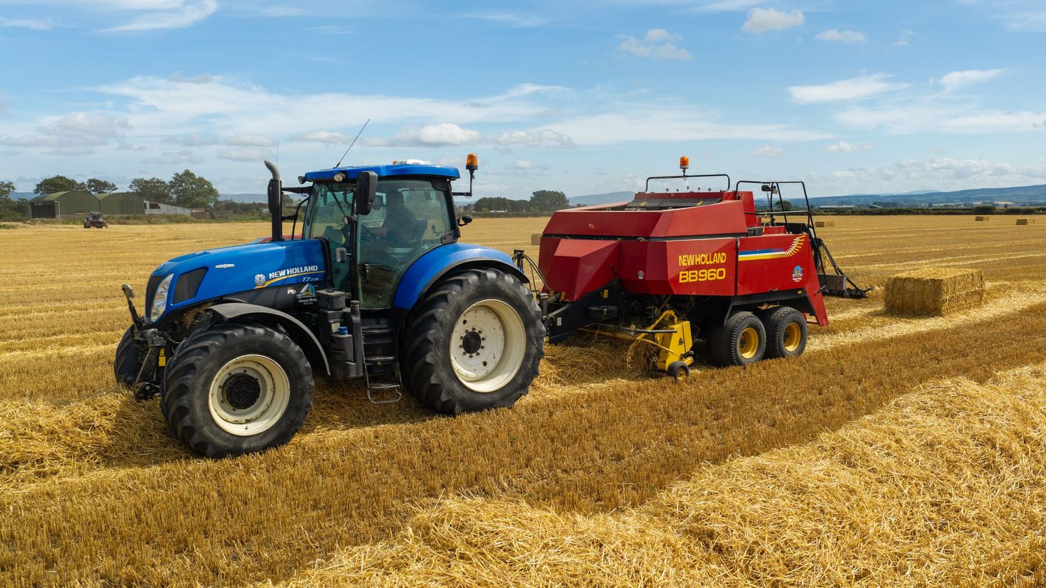 Blue New Holland tractor pulling a red square baler harvesting straw bales in a golden field.