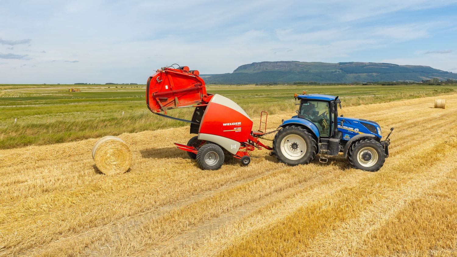 Blue New Holland tractor with red Lely round baler harvesting hay bales in a golden field with mountains in background.