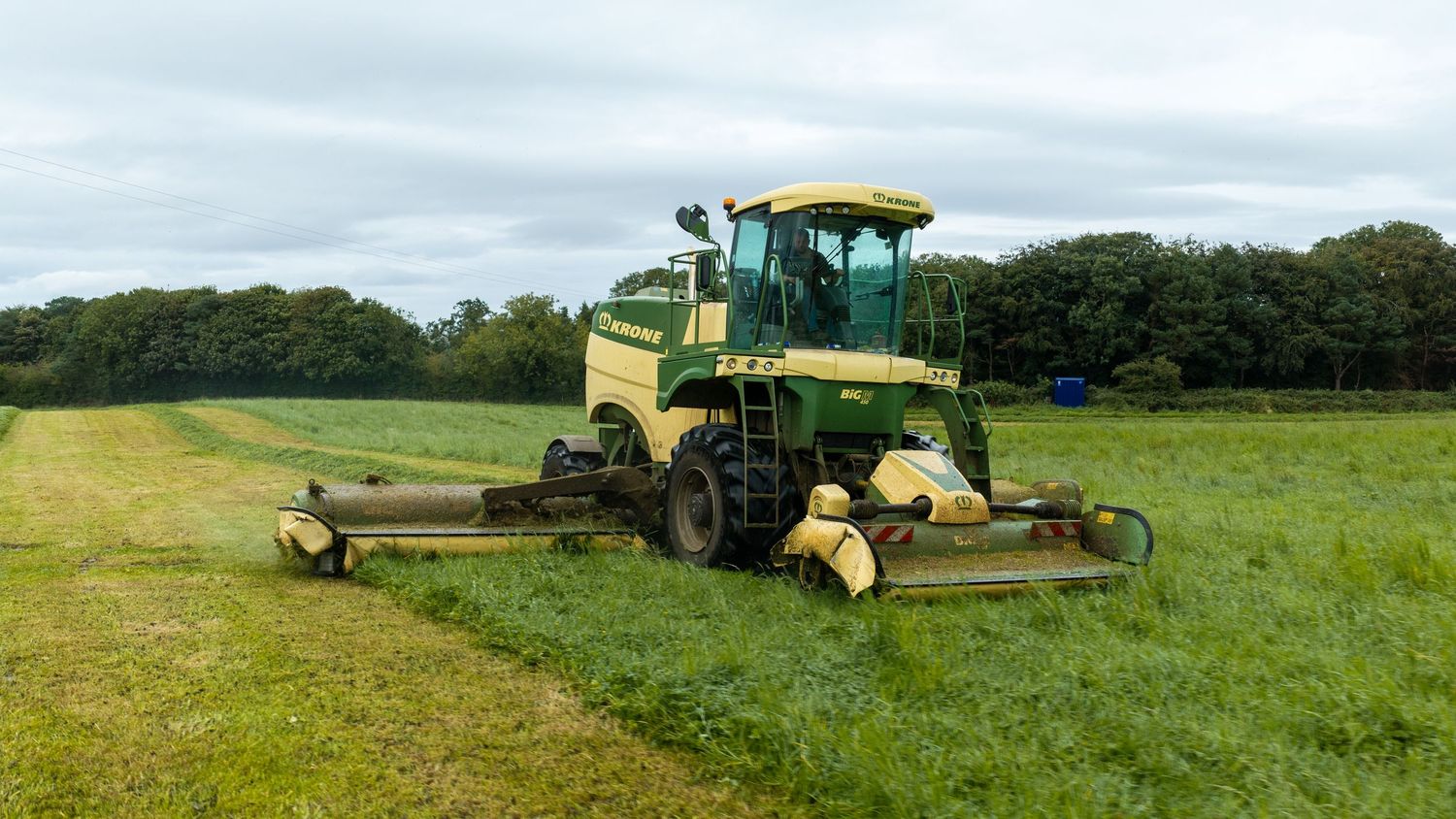 Green John Deere combine harvester mowing a lush grass field with tree line in background.