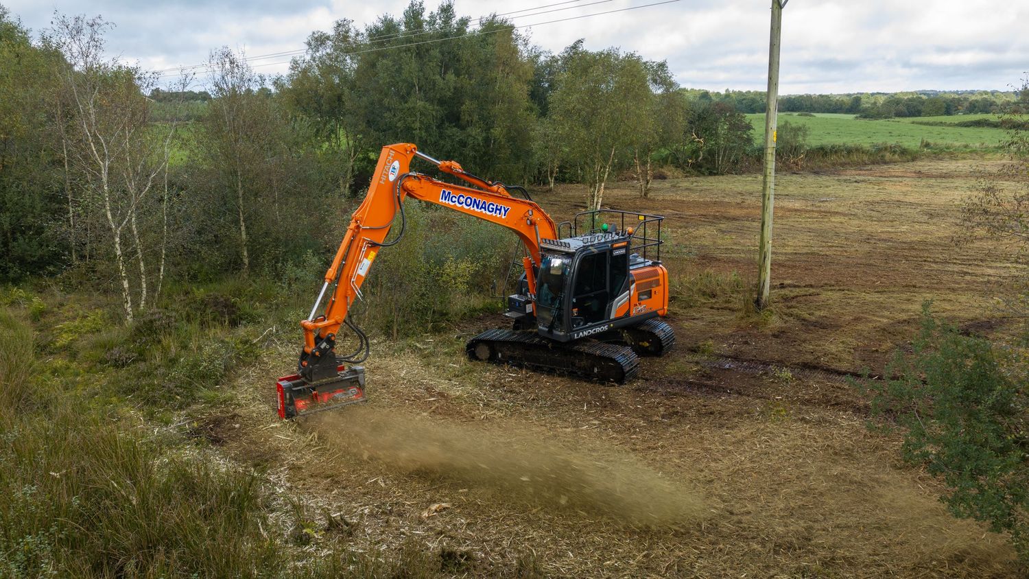 Orange Doosan excavator clearing land with a mulcher attachment in a rural field surrounded by trees.