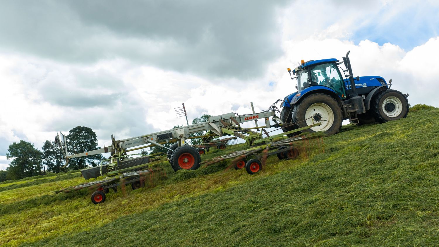 Blue New Holland tractor pulling farm equipment on a green hillside under cloudy skies.