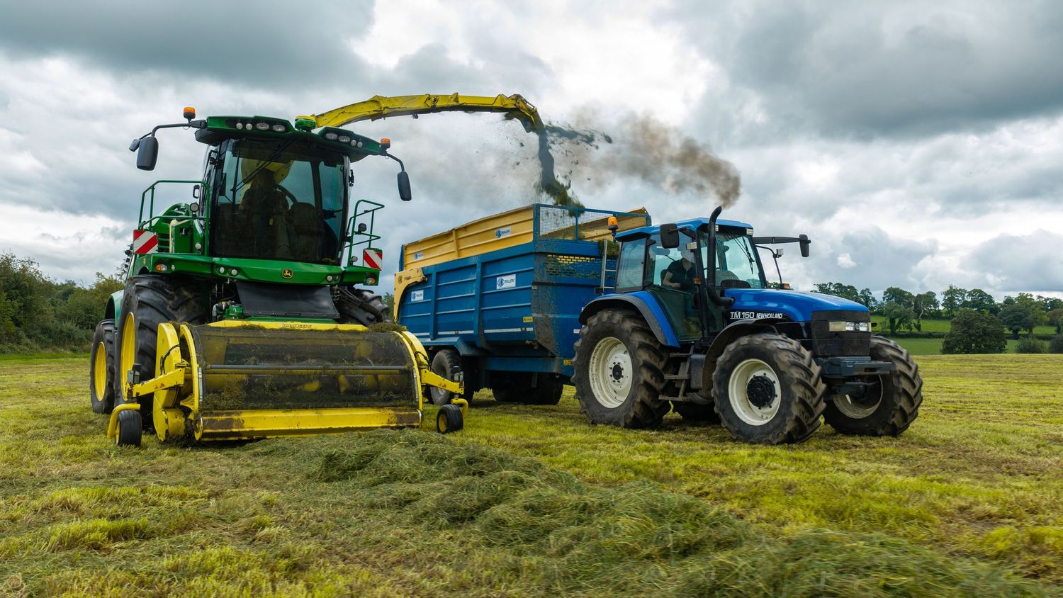 Green John Deere forage harvester and blue New Holland tractor working together in a lush farm field.