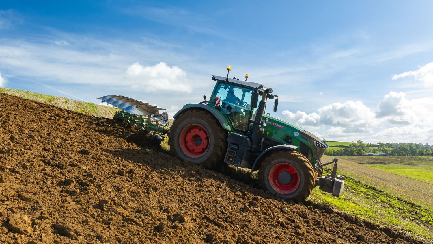 Green tractor with red wheels plowing a brown field on a hillside under a partly cloudy blue sky.