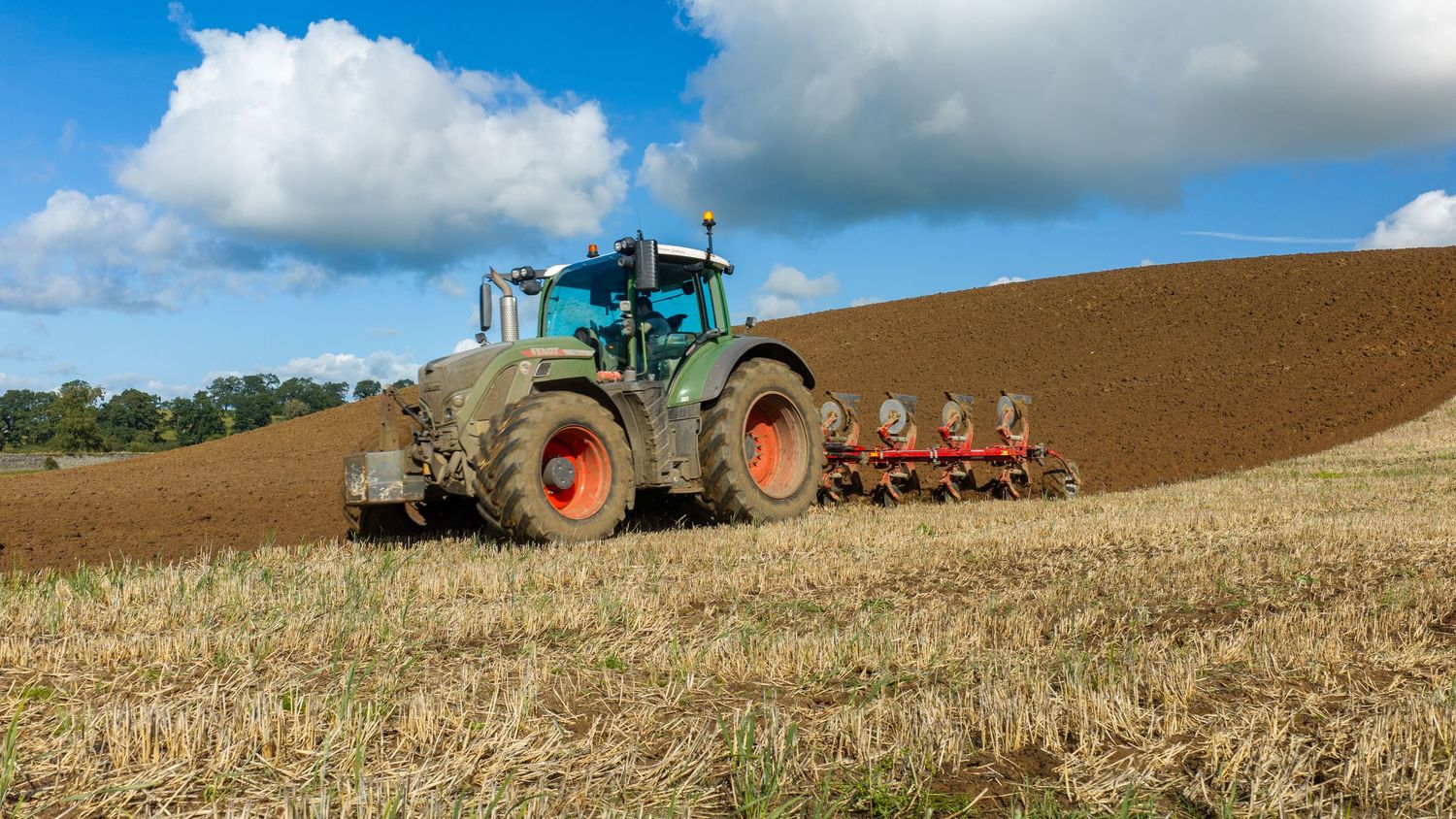 Green tractor plowing a brown agricultural field under a blue sky with white clouds on a sunny day.