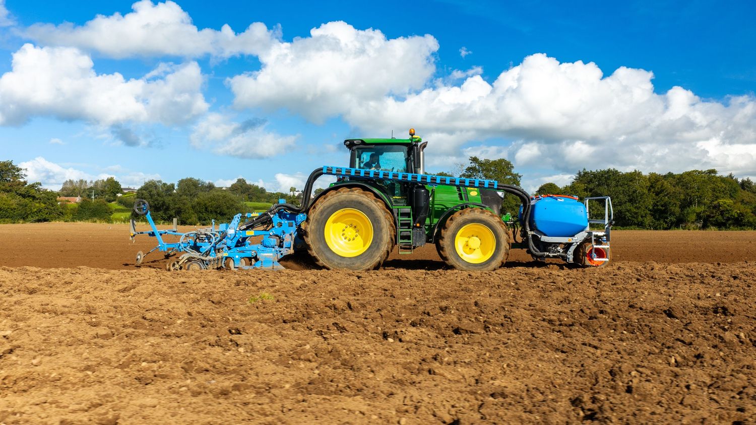 Green John Deere tractor plowing a brown agricultural field under a blue sky with white clouds.