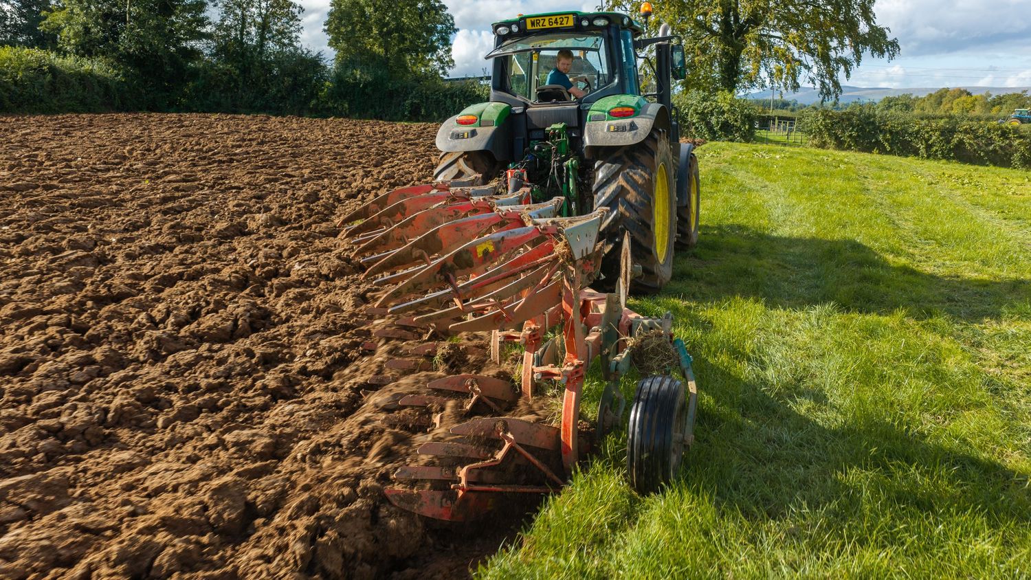 Green tractor plowing a brown agricultural field with a red multi-furrow plow on a sunny day.