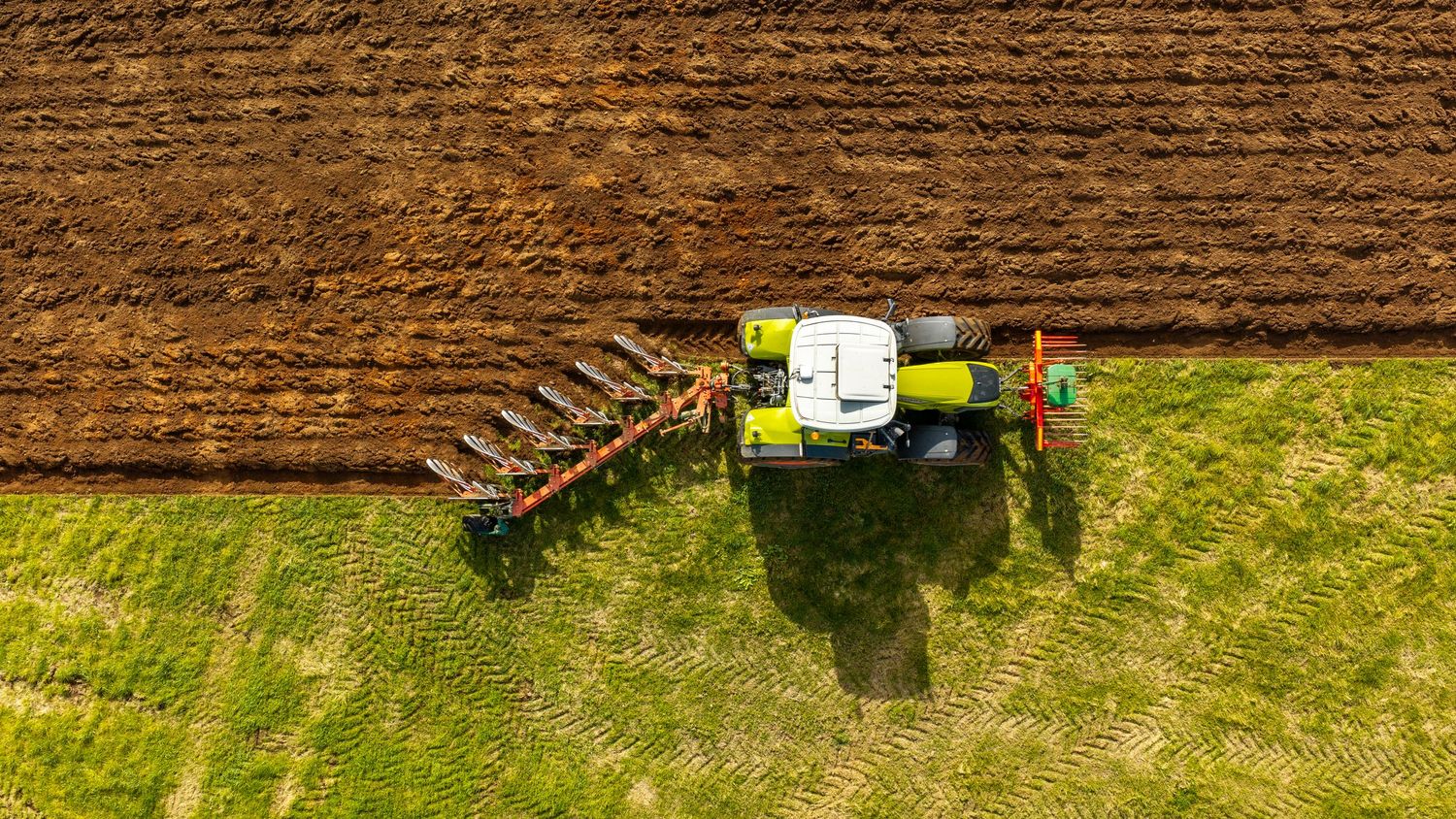 Aerial view of a green CLAAS tractor with red cultivator attachment plowing a large agricultural field.