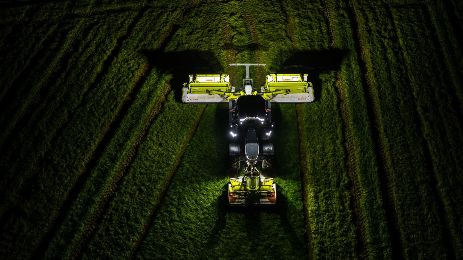 Aerial night view of a illuminated green tractor with wide harvesting attachments working in a dark field.