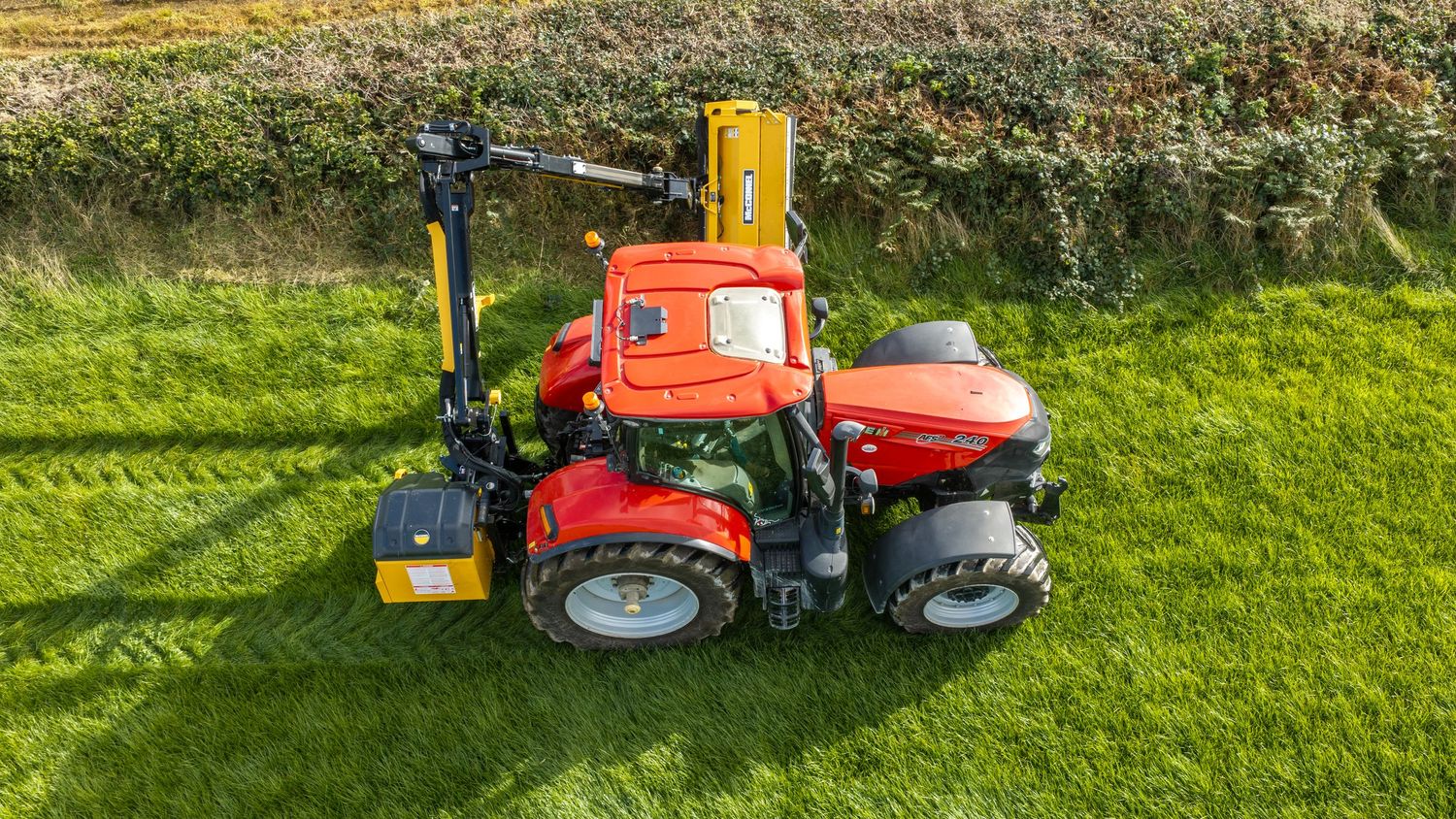 Red Case IH tractor with hedge cutter attachment trimming green hedgerow, aerial view on lush grass field.