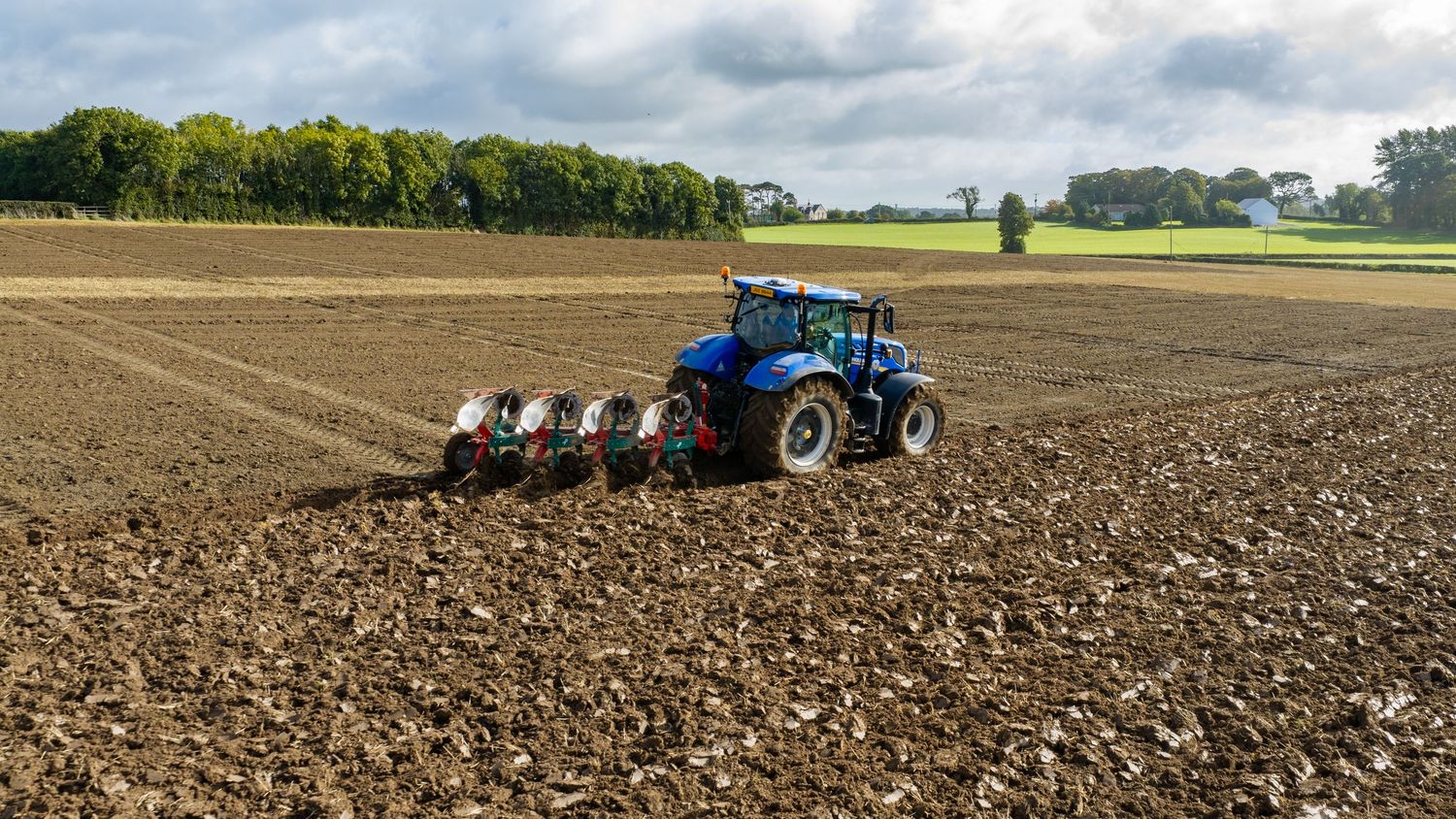 Blue New Holland tractor plowing a large agricultural field under a cloudy sky in autumn.