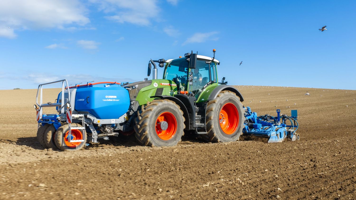 Green Fendt tractor with blue tank attachment plowing a dry field under a clear blue sky.