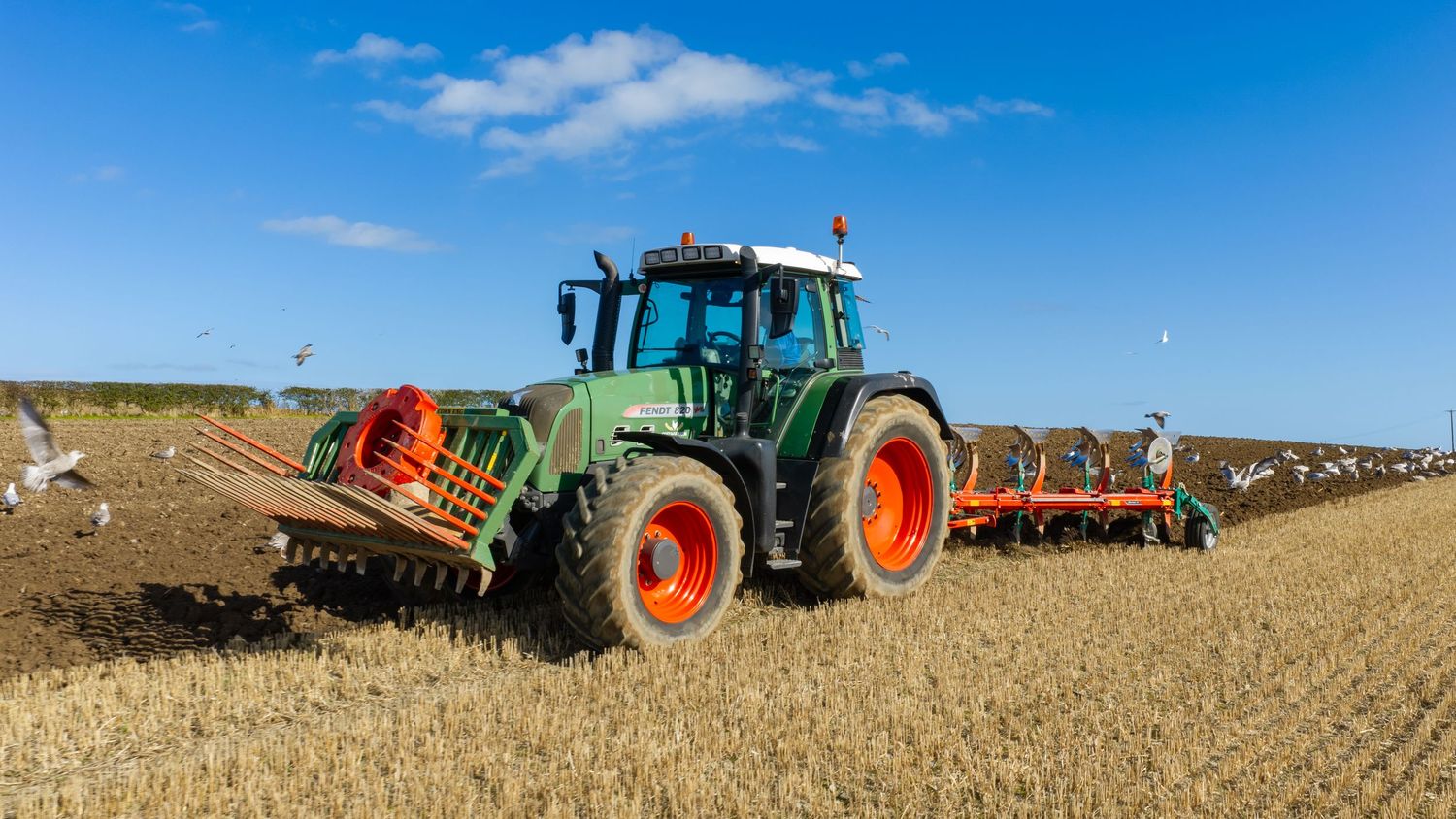 Green tractor with orange wheels plowing a field under a blue sky on a sunny day.