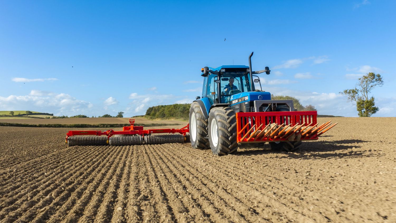 Blue tractor with red seeder attachment cultivating a large agricultural field under a bright blue sky.