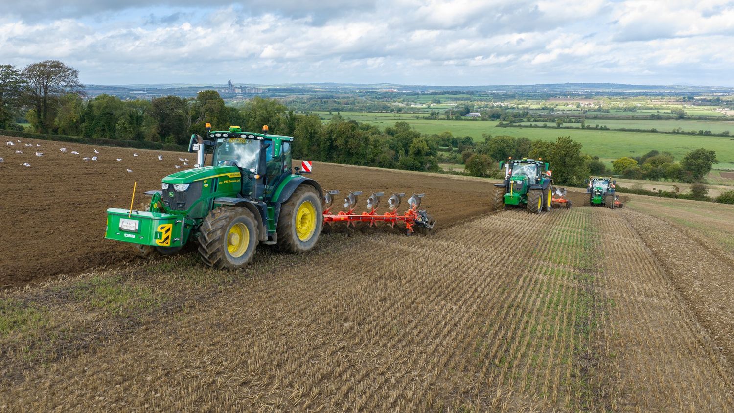 Two John Deere tractors plowing a large agricultural field with rolling green hills in the background.
