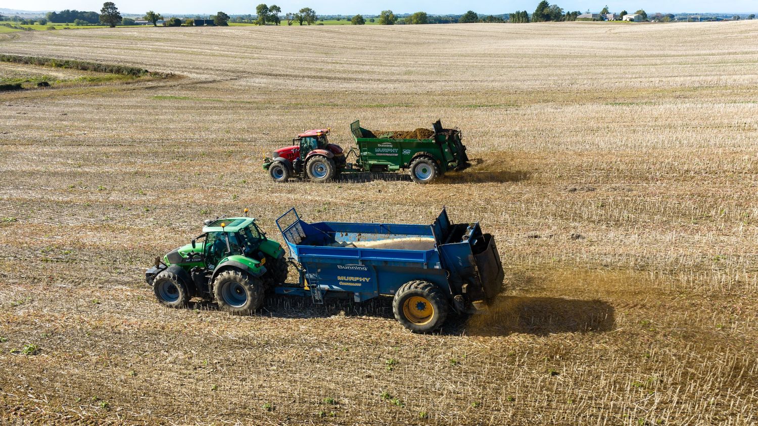 Two tractors harvesting golden wheat fields on a sunny day, with farm equipment trailing behind in rural farmland.