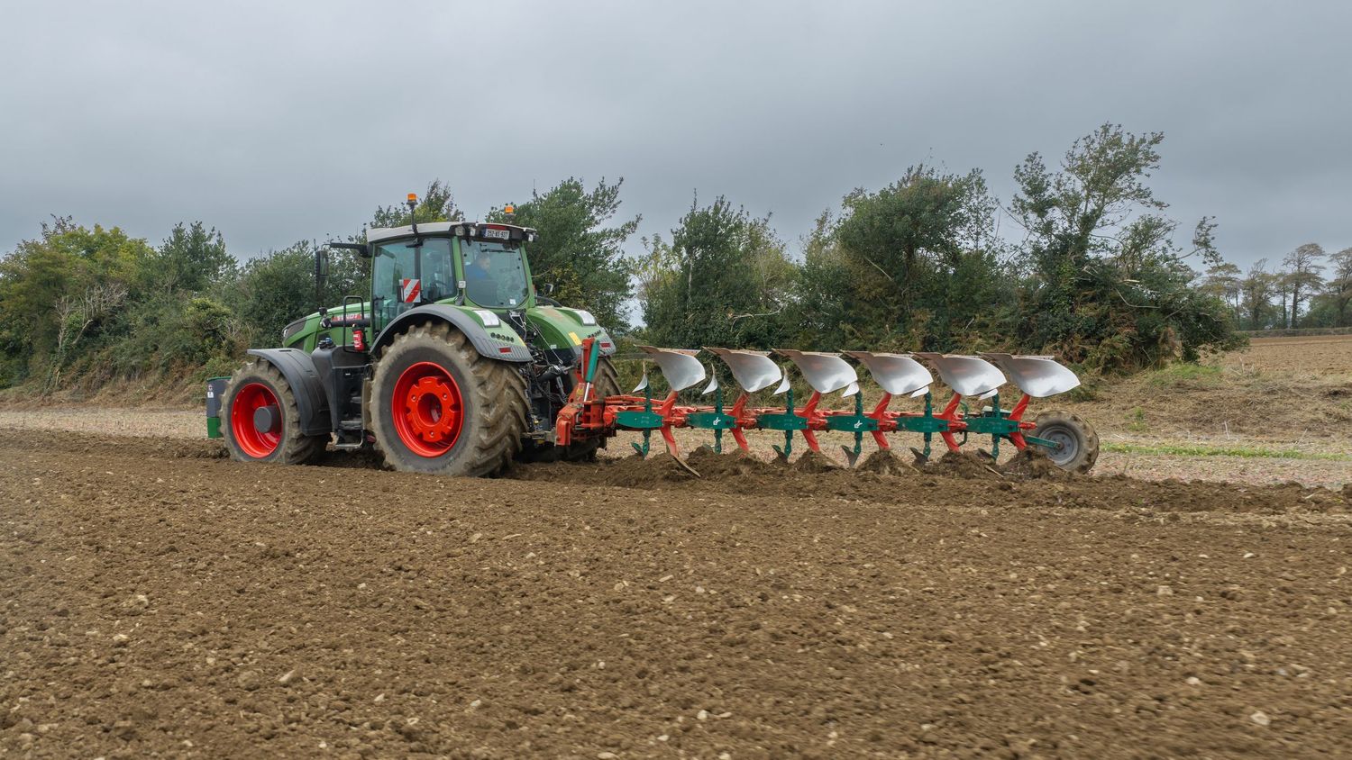 Green Fendt tractor with multi-bottom plow attachment tilling a large agricultural field on an overcast day.