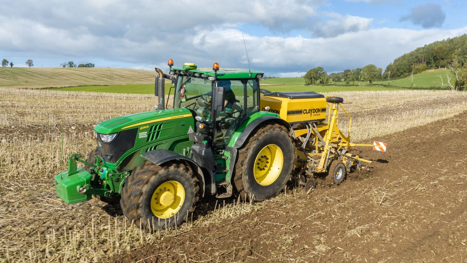 Green John Deere tractor pulling a seed drill across a freshly plowed agricultural field under a clear blue sky.