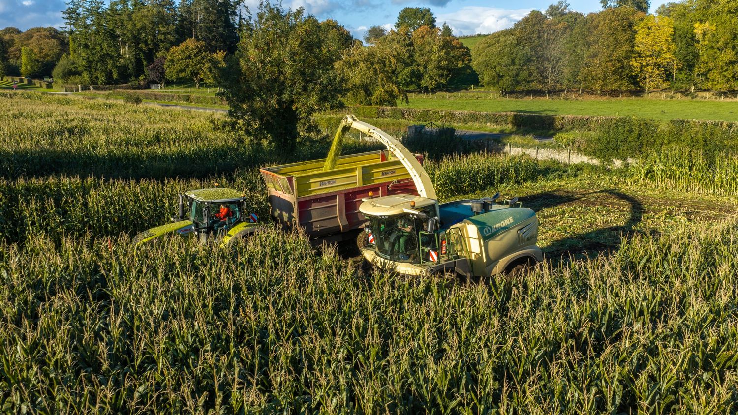 Aerial view of green farm machinery harvesting crops in a lush field surrounded by trees on a sunny day.