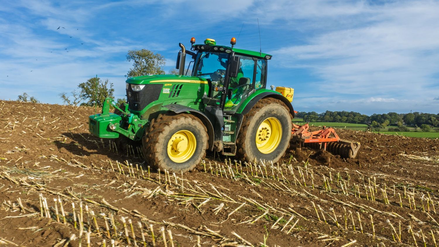 Green John Deere tractor plowing a field on a sunny day with a red tillage implement attached.
