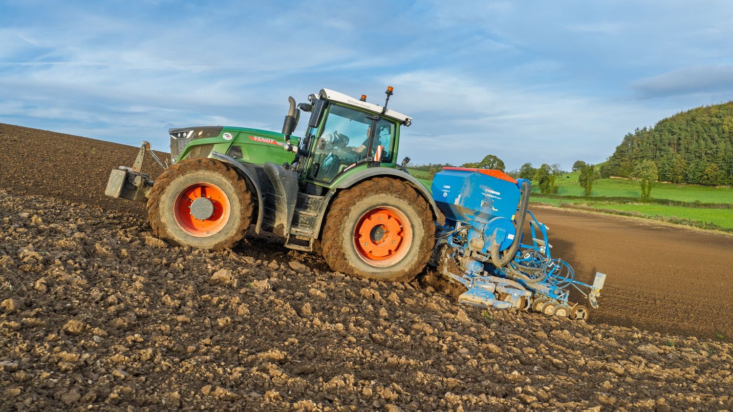 Green tractor with orange wheels plowing a large agricultural field under a blue sky.