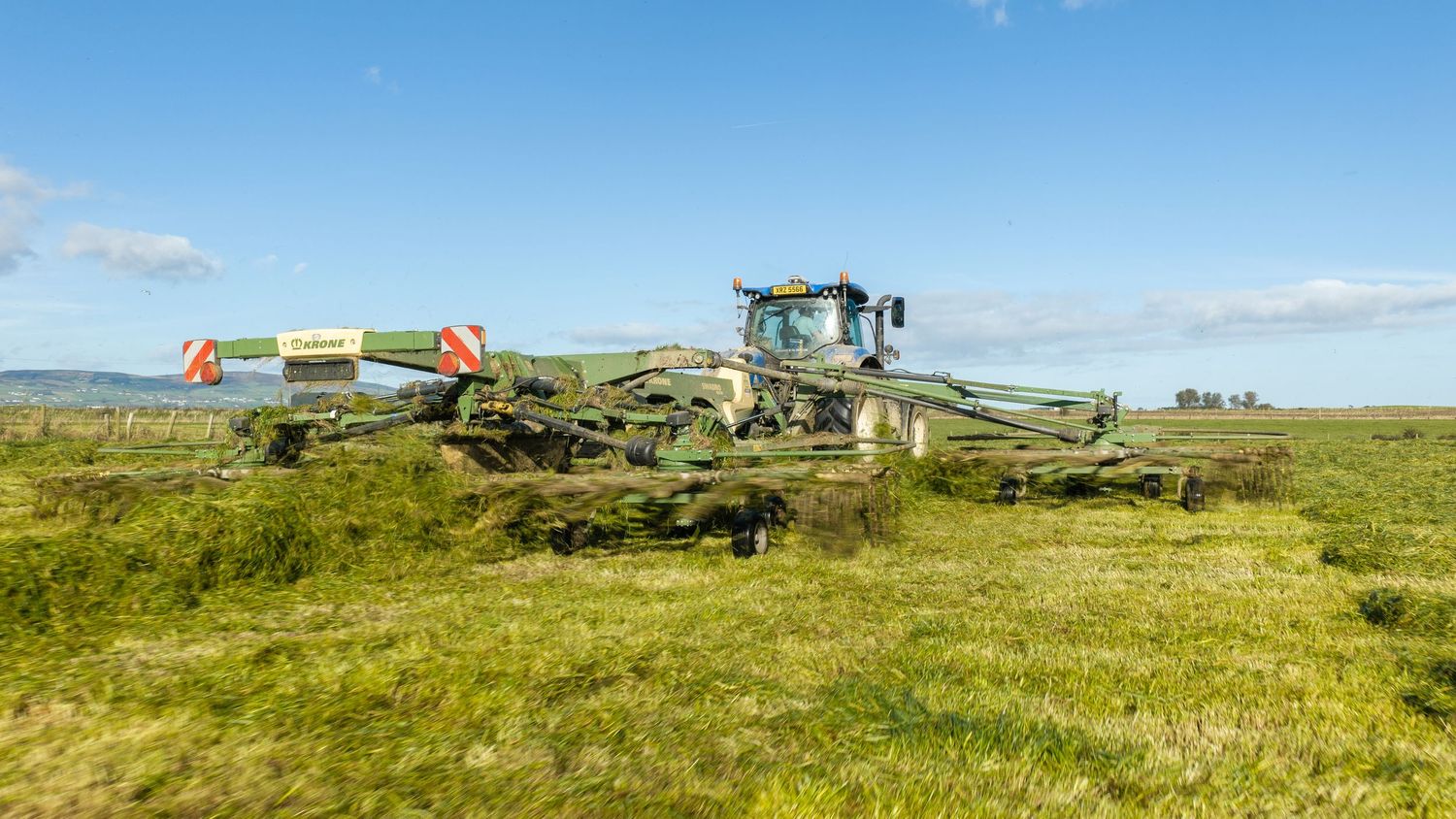 Tractor pulling a wide green hay rake across a grassy field under a blue sky with scattered clouds.