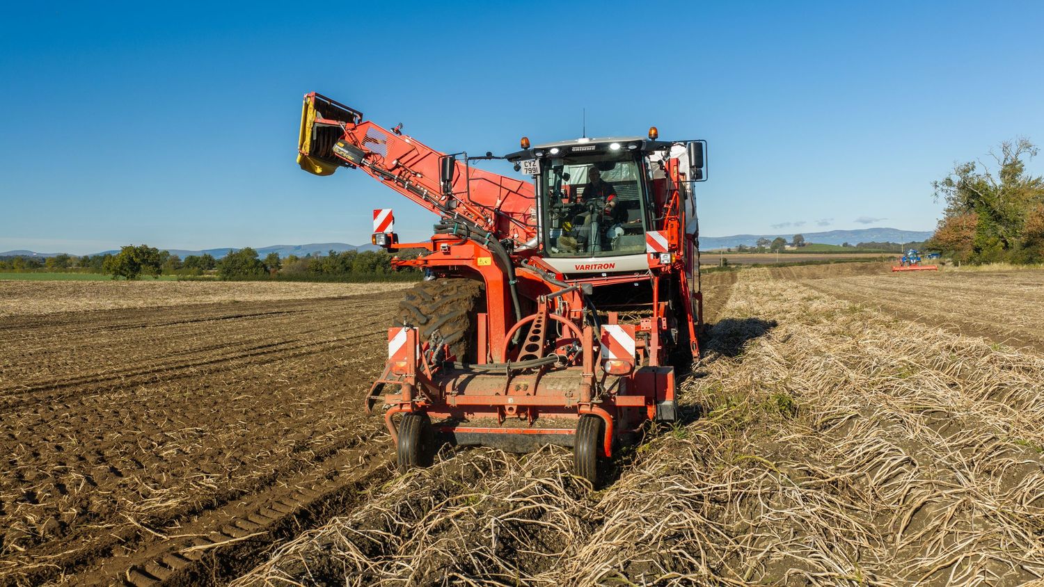 Orange root crop harvester machine driving across a plowed farm field under a clear blue sky during harvest season.