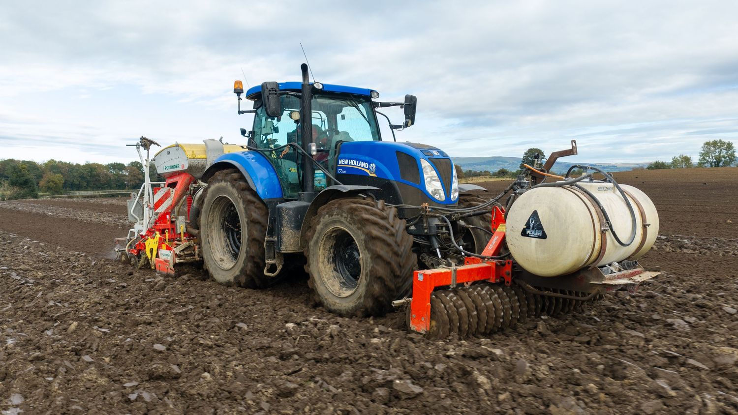 Blue tractor pulling a seed drill and tank across freshly tilled brown soil in a farm field under a partly cloudy sky.