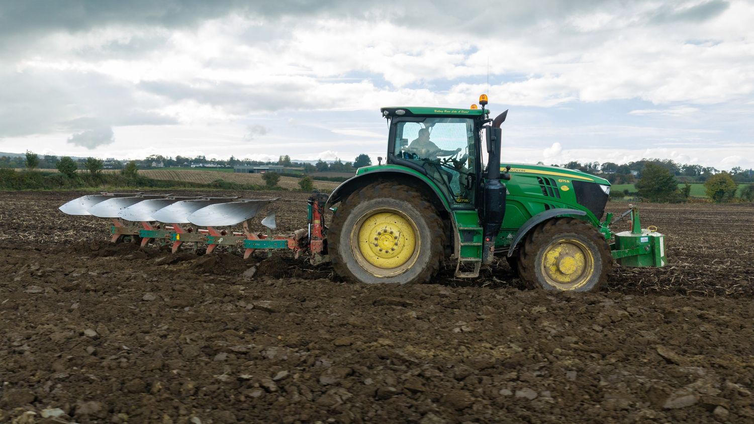 Green John Deere tractor pulling a multi-furrow plough across dark, freshly turned soil in a farm field under an overcast sky.
