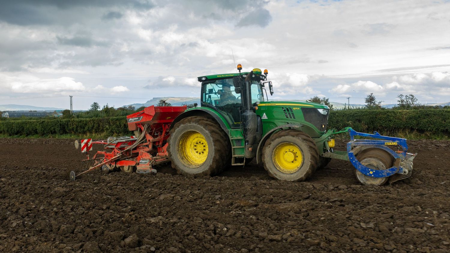 Green tractor with yellow wheels pulling a red seed drill across freshly tilled farmland under a cloudy sky.