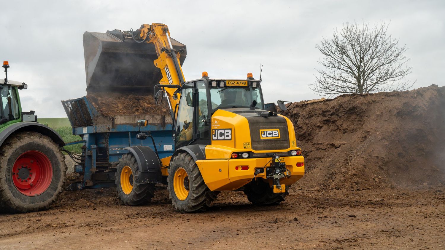 JCB wheeled excavator loading dirt into a blue trailer on a construction site with a green tractor nearby.