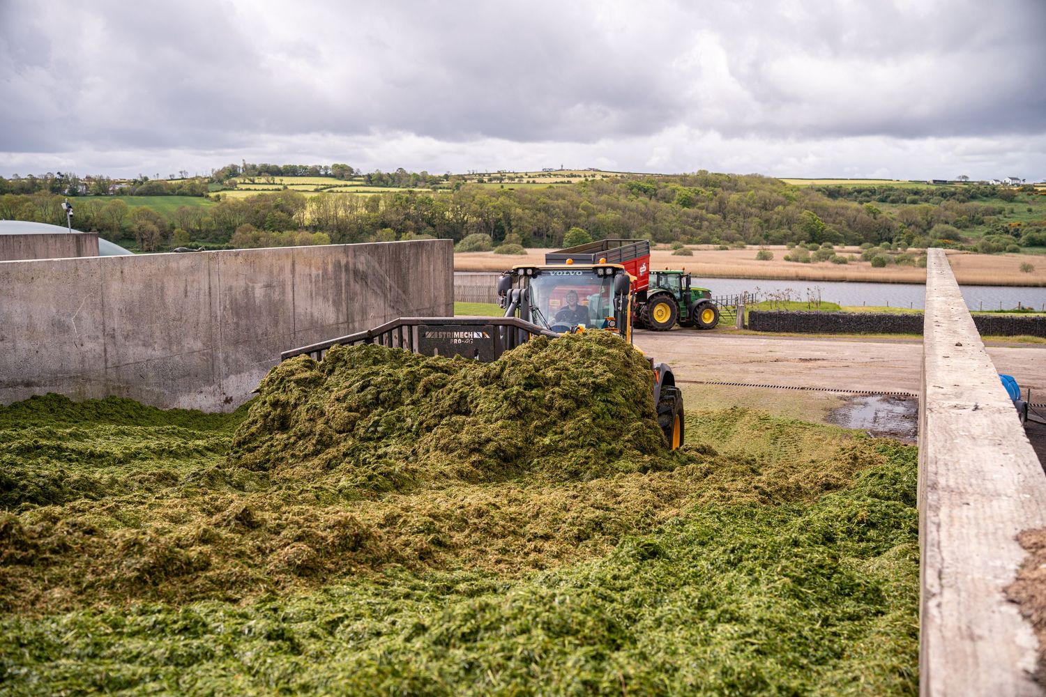 Green tractor compacting silage on a farm clamp with concrete walls, surrounded by rural countryside landscape.