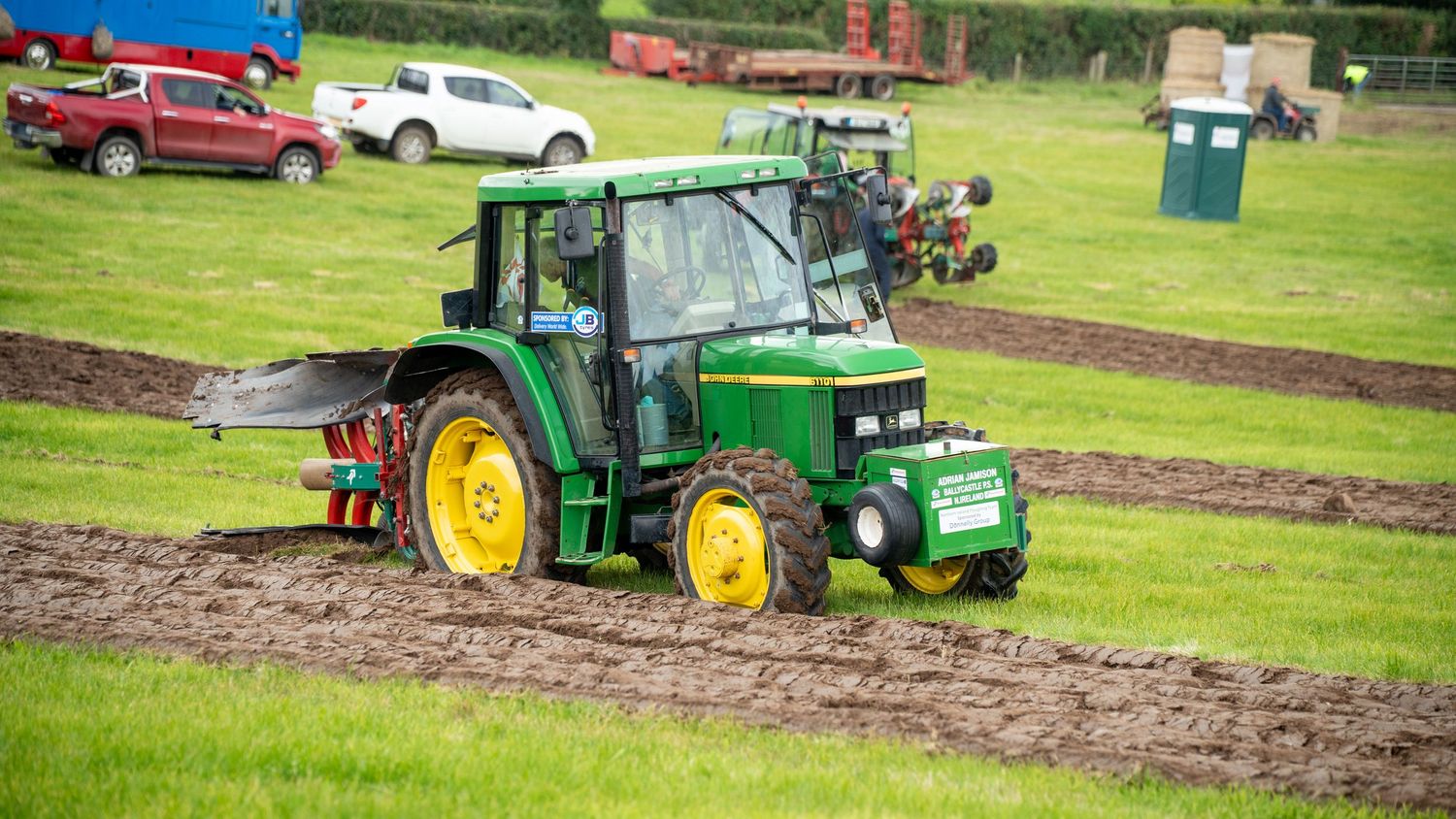 Green John Deere tractor with yellow wheels working muddy agricultural field during outdoor farming event.