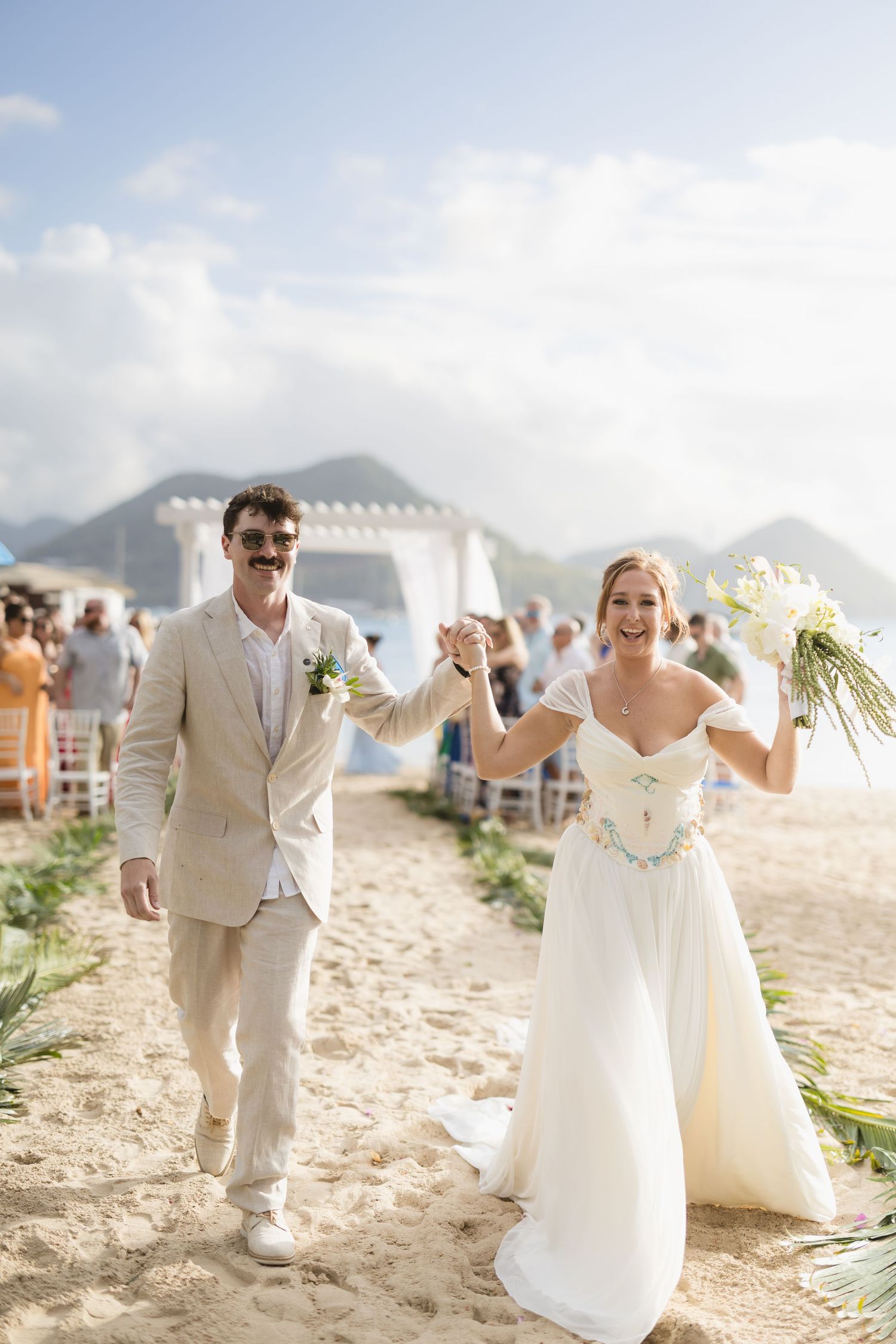 A couple celebrates during their beach wedding ceremony with mountains and a gazebo in the background.