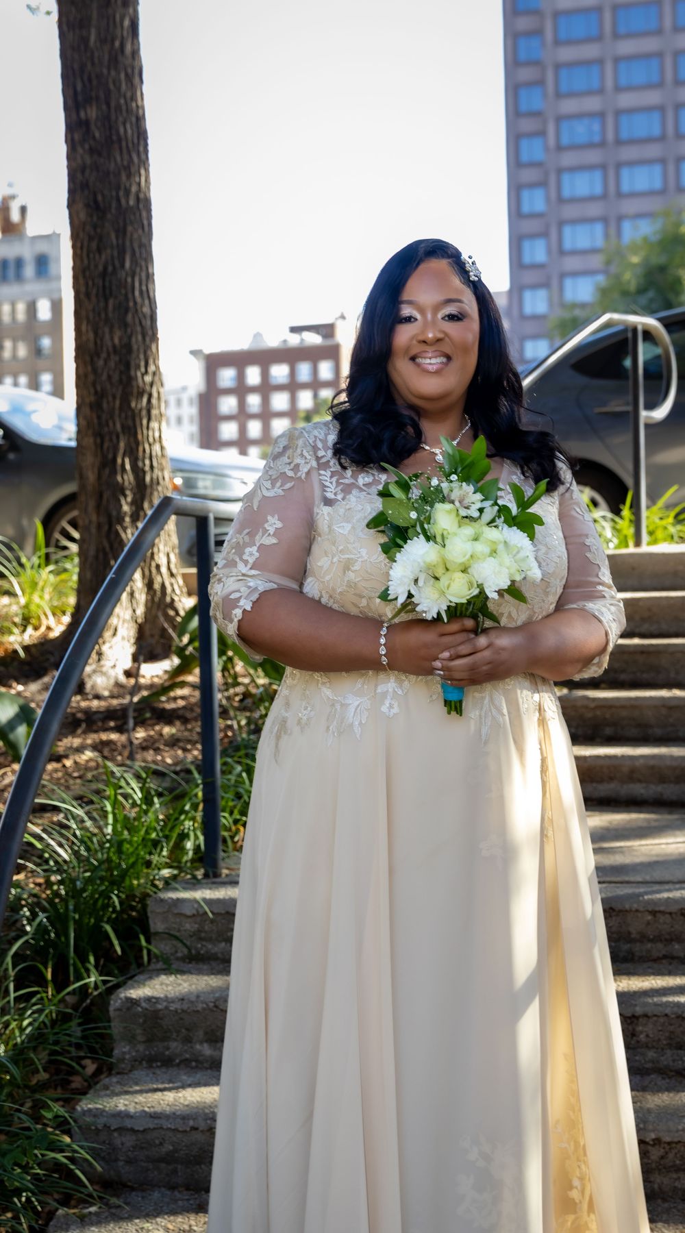 A bride in a flowing cream-colored gown holds a white floral bouquet in an outdoor garden setting.