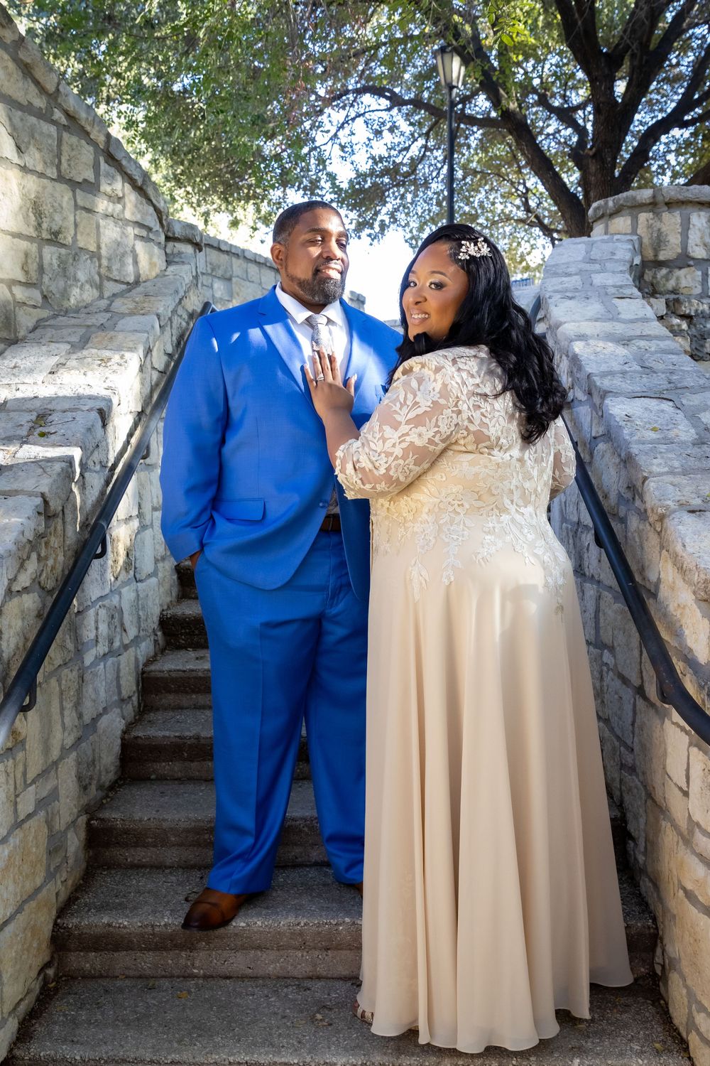 A couple in formal attire poses together on stone steps, wearing a royal blue suit and champagne-colored gown.
