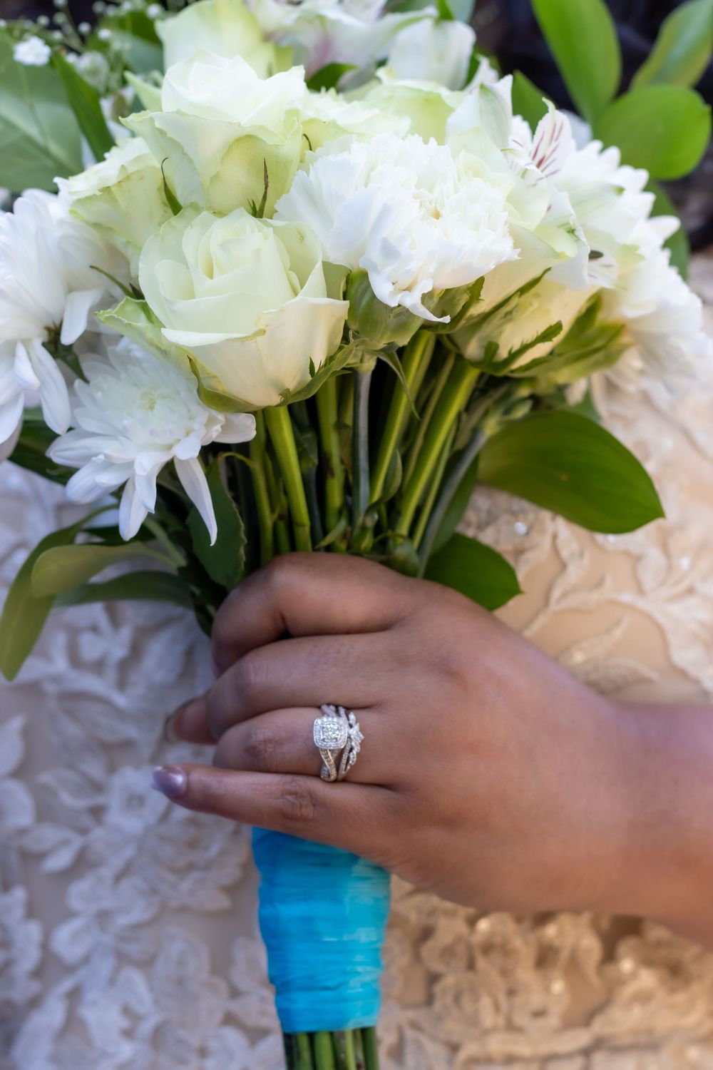 A hand wearing an engagement ring holds a white flower bouquet wrapped in turquoise ribbon.