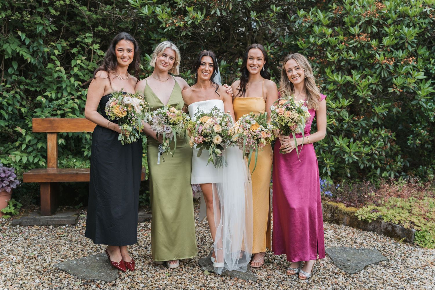 A wedding party in mismatched silk dresses stands together in a garden setting holding floral bouquets.