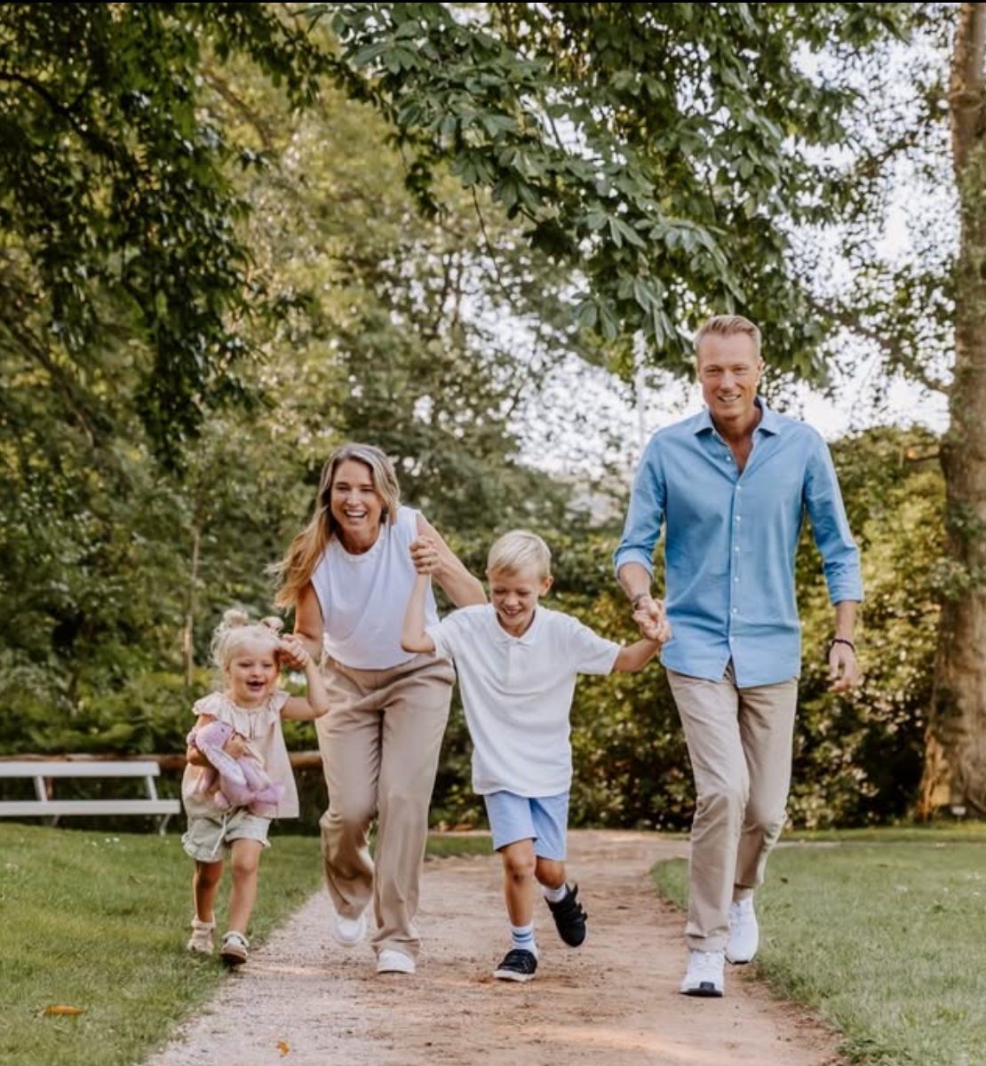 En familie i koordinerede outfits løber og leger sammen på en jordsti omgivet af grønne træer på en solrig dag. Fotograf Middelfart