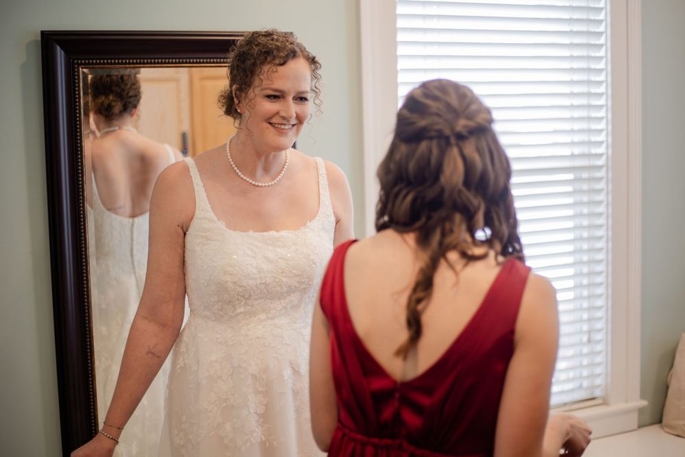A bride and bridesmaid in red dress help with wedding preparations near a bright window.