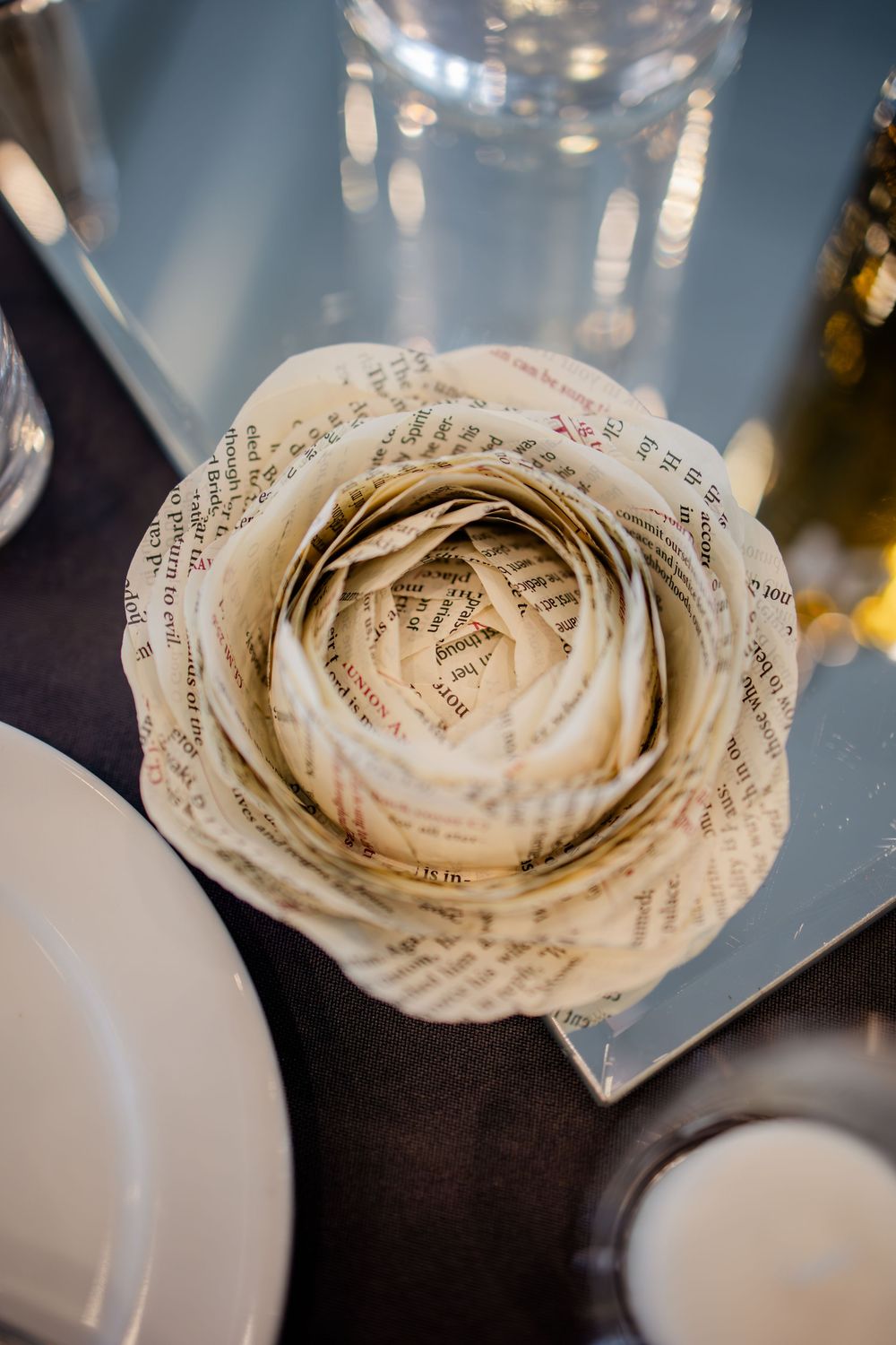 A delicate paper flower crafted from vintage book pages sits on a reflective surface next to white dishes.