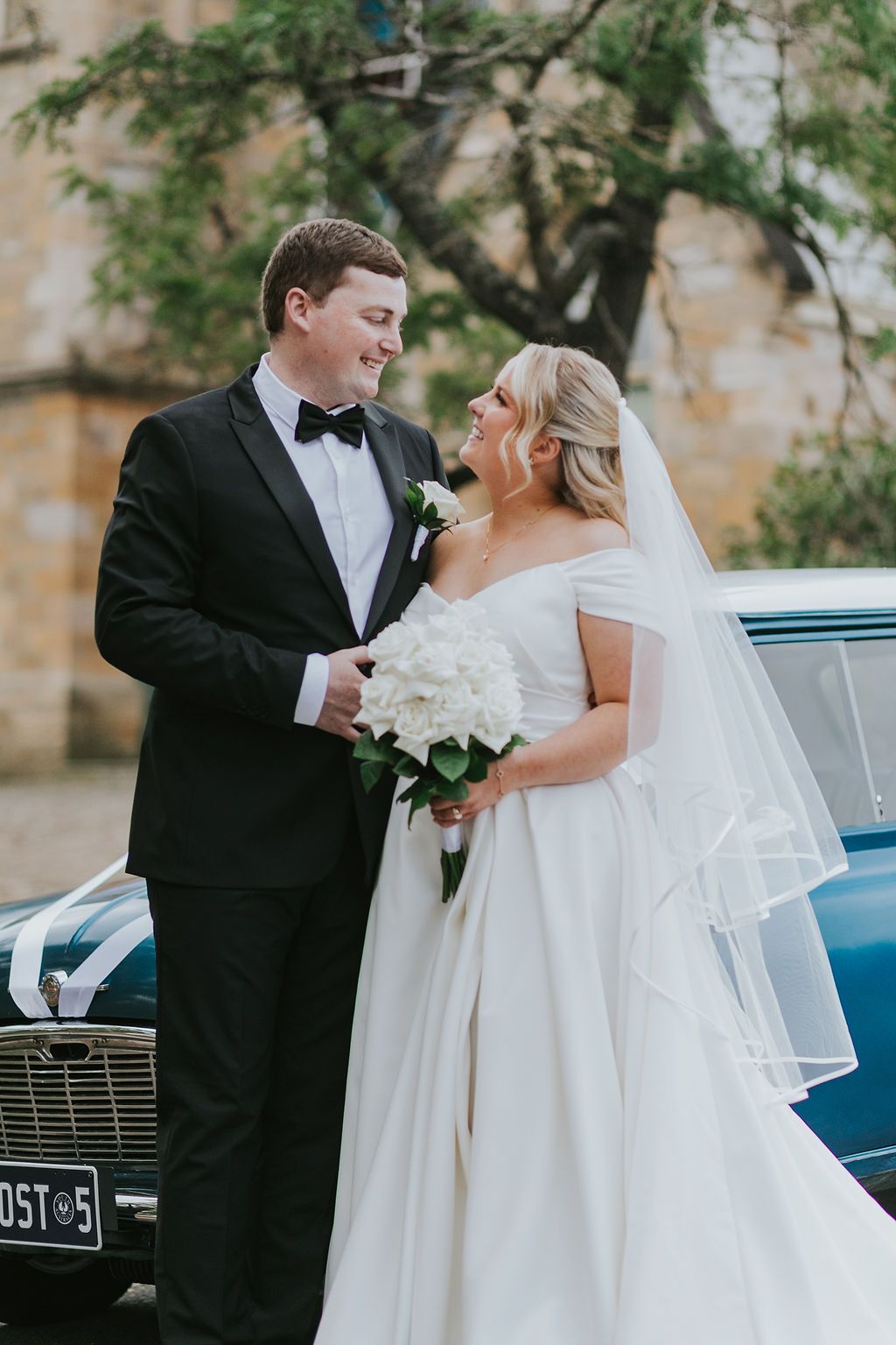 Bride and groom share a romantic moment beside a vintage blue car, bride holding white bouquet in elegant off-shoulder gown.