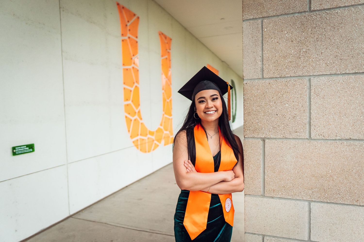 Graduate in cap and gown poses confidently against wall with orange university logo. UTD Graduate Portraits by Eric Aaberg Photography in DF
