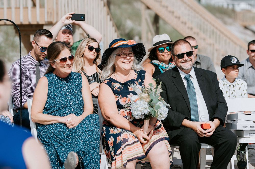 Wedding guests seated outdoors in formal attire watching ceremony proceedings.