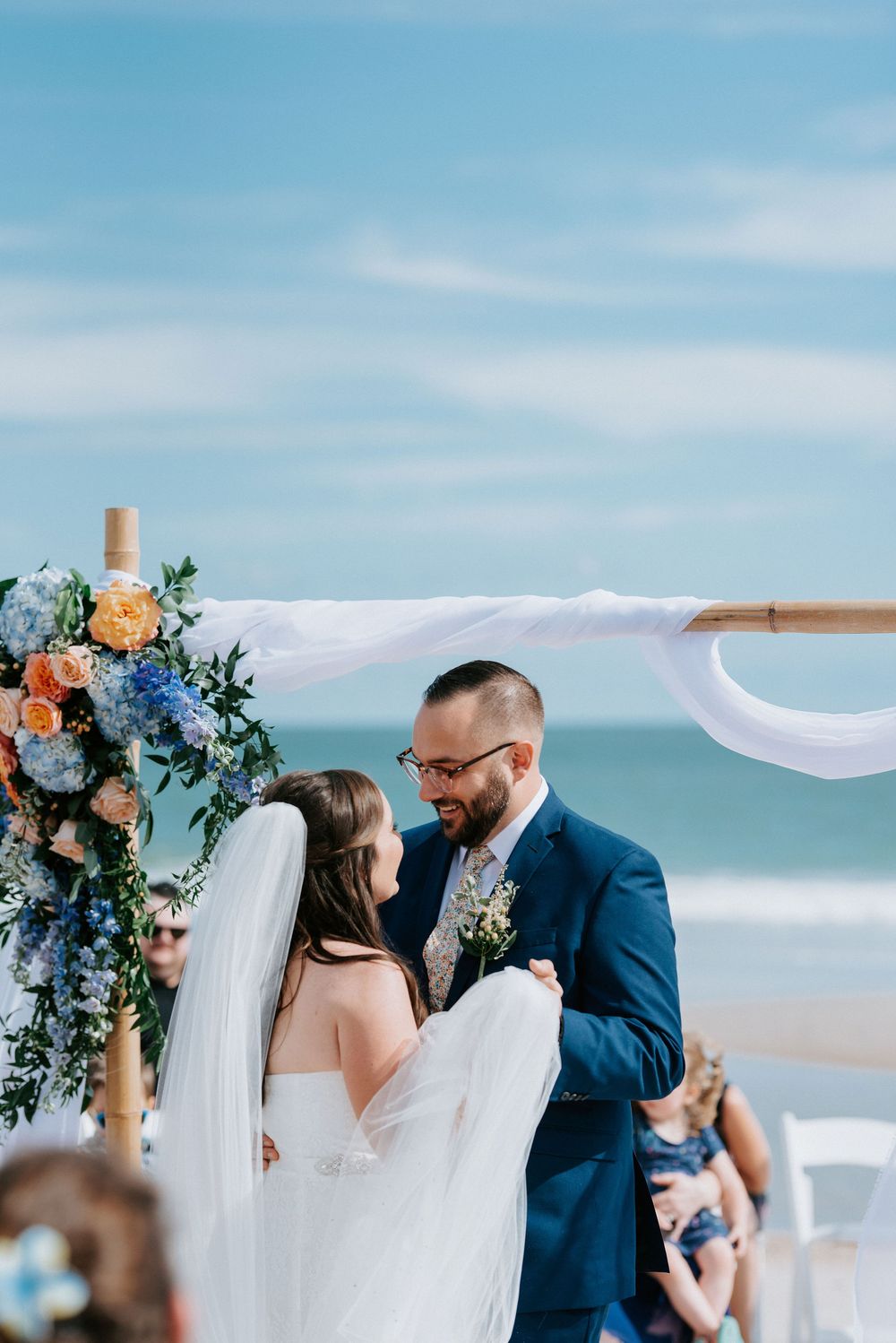 Beach wedding ceremony setup with white fabric arch and blue and peach floral arrangements by the ocean.
