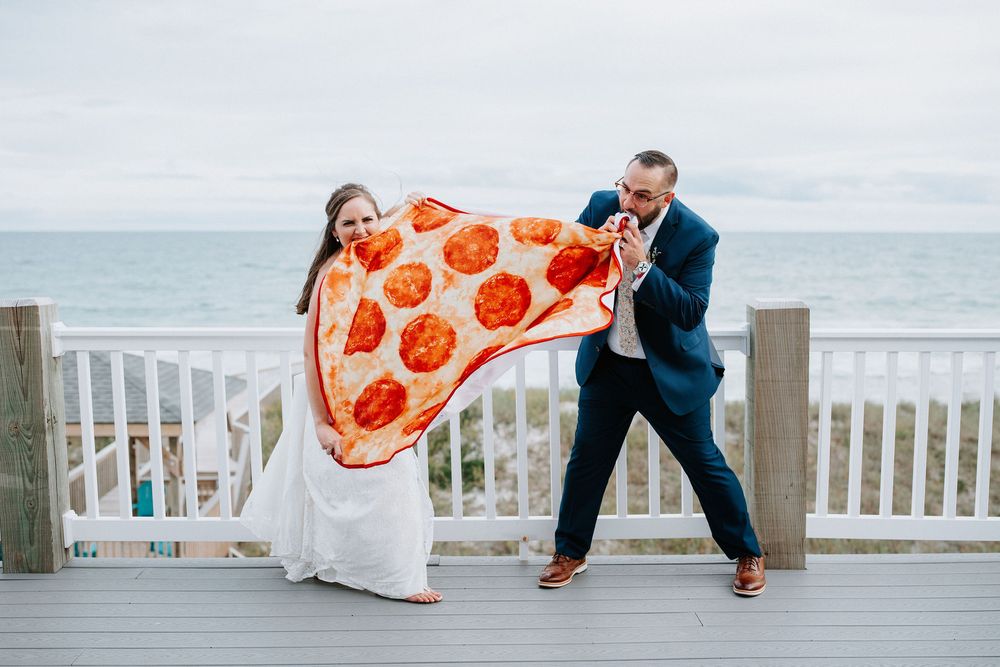 Playful newlyweds posing with pizza slice props on a beachfront deck during their wedding photoshoot.