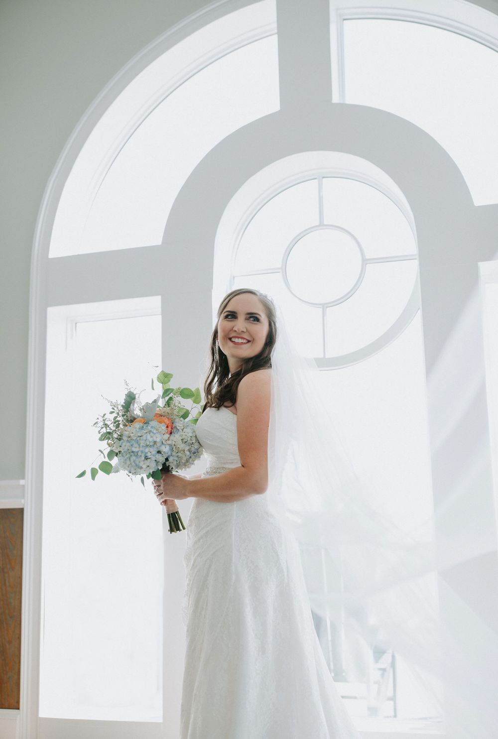 A bride in a white wedding dress holds a bouquet while standing in a bright room with arched windows.