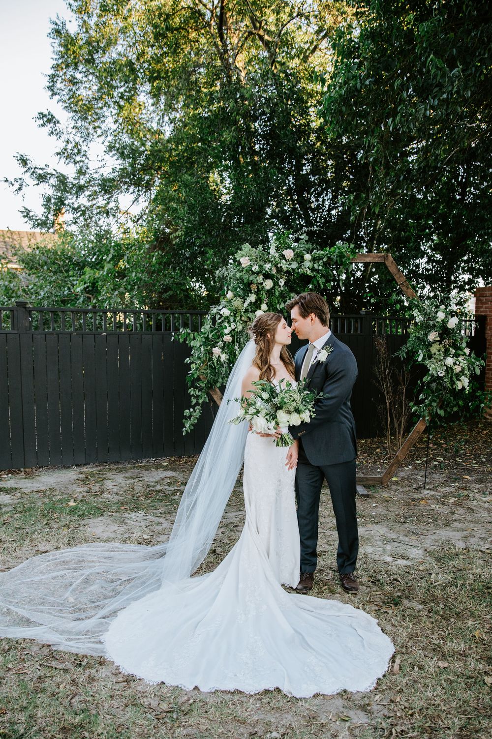 A couple shares a romantic moment in a garden setting wearing wedding attire with a flowing white dress train.