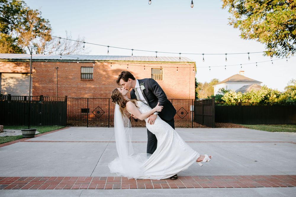 A couple in wedding attire dances and twirls across a brick courtyard with string lights overhead at sunset.