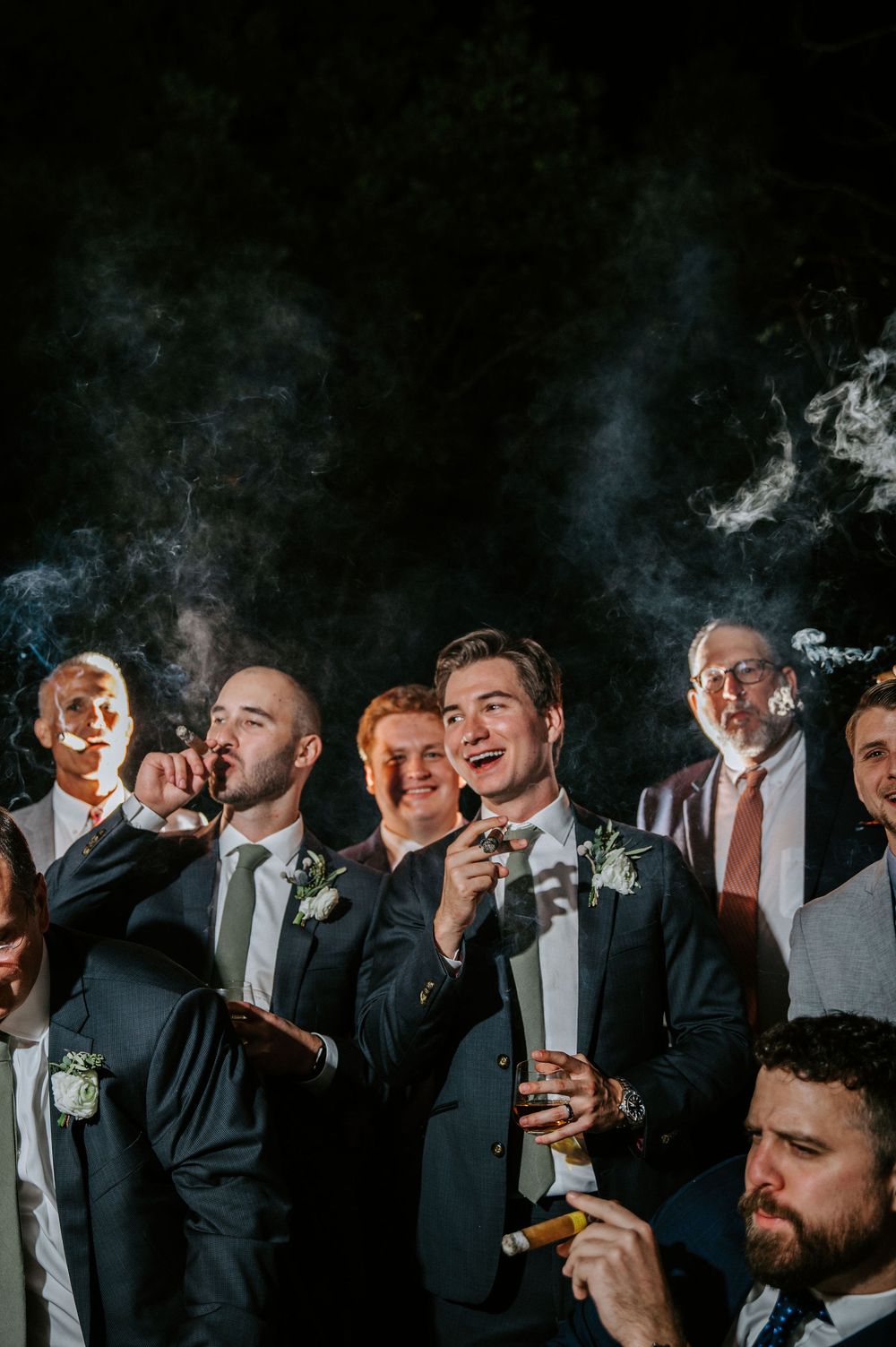 Groomsmen enjoying cigars and drinks in dramatic black and white nighttime photography.