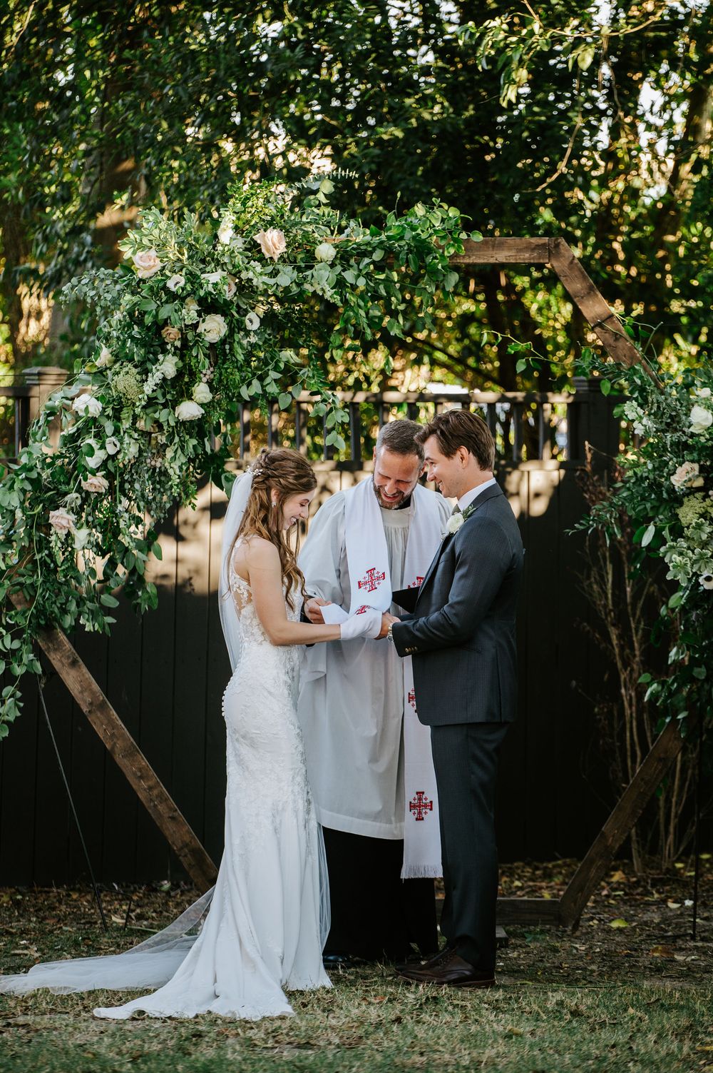 A wedding ceremony takes place under a beautiful floral arch with white roses and greenery in an outdoor garden setting.